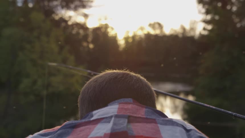 A boy sits in front of a river while someone near him is holding a fishing pole