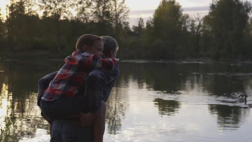 Father gives son a piggyback ride while standing on lake shore and looking across to trees on opposite shore
