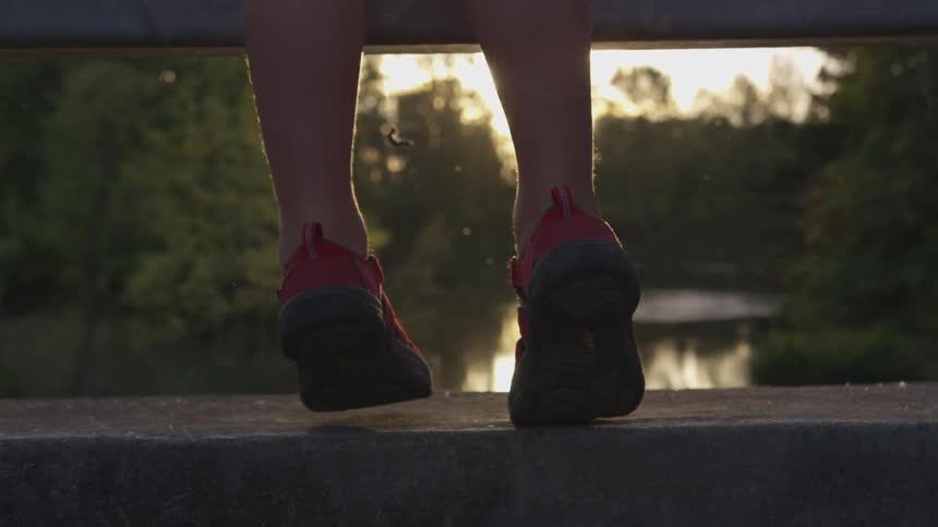 Person wearing red shoes is standing on tiptoe atop a bridge railing overlooking a lake surrounded by forest trees