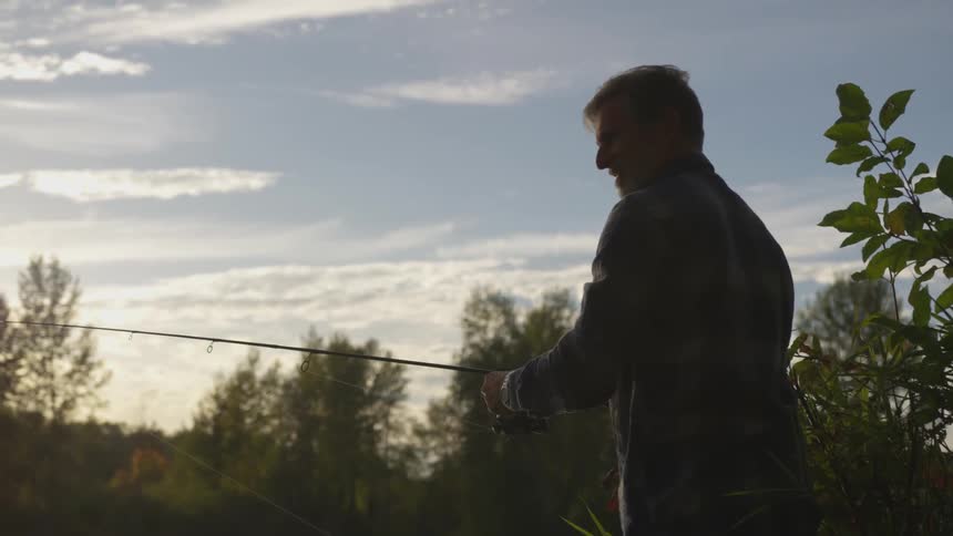 An man in a flannel turns the reel of his fishing rod while surrounded by plants and trees