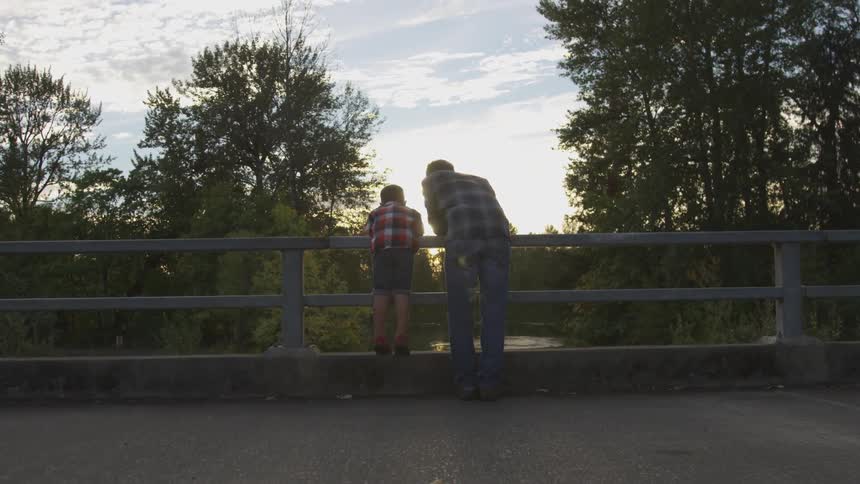 A dad leans on a fence next to his son as they both look down at the river