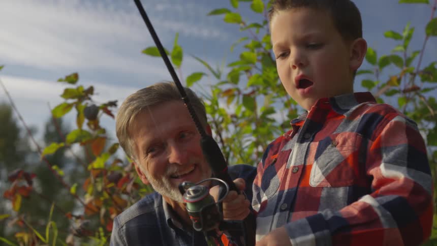 A grandfather is encouraging his grandson who is trying to fish with a real fishing rod and reel