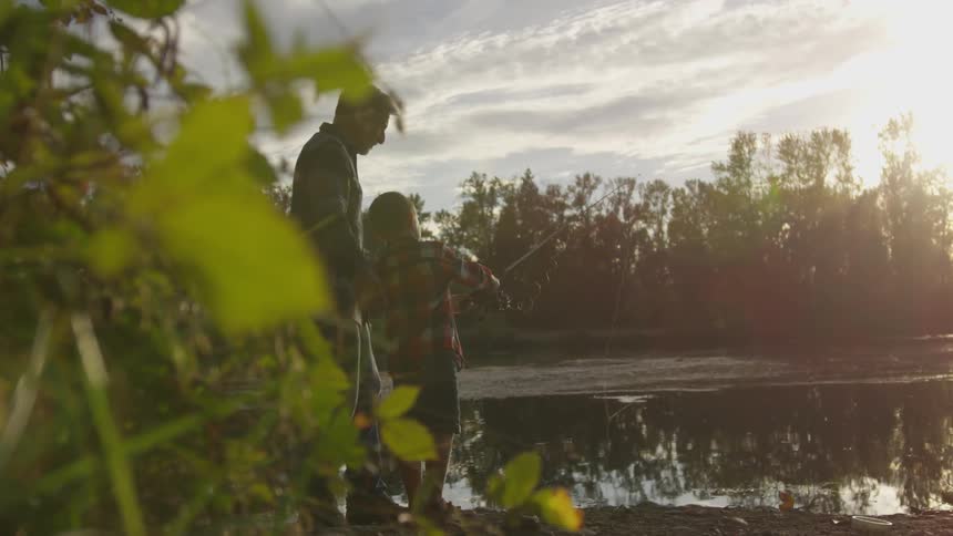 An older man helps a young boy cast his fishing line into a calm river