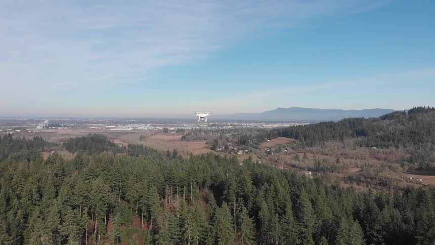 A white drone with multiple propellers ascends above a dense green forest