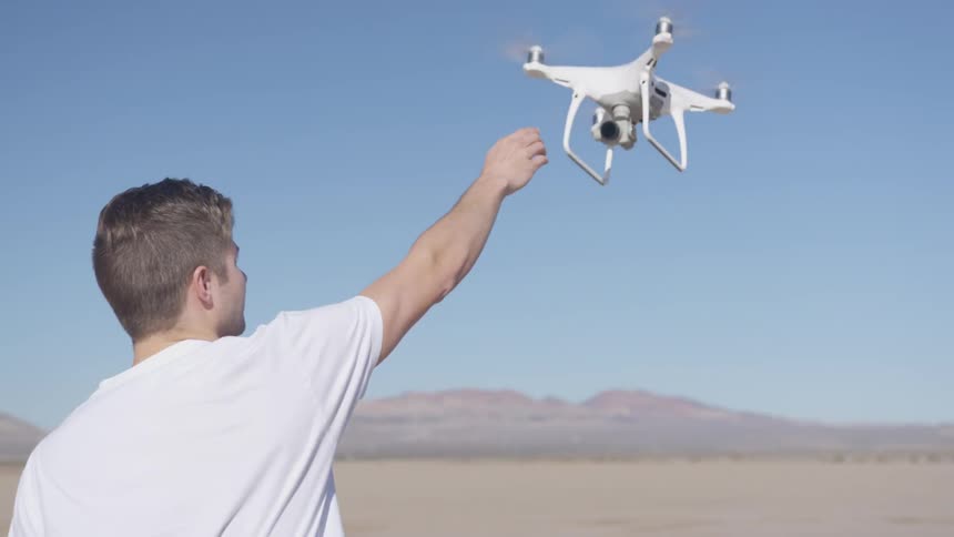 A man in a white tee shirt reaches up to grab a quadcopter drone that he was flying