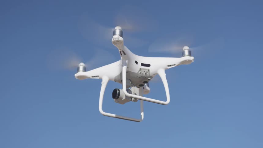 A white drone with propellers spinning is hovering in one spot in the blue sky