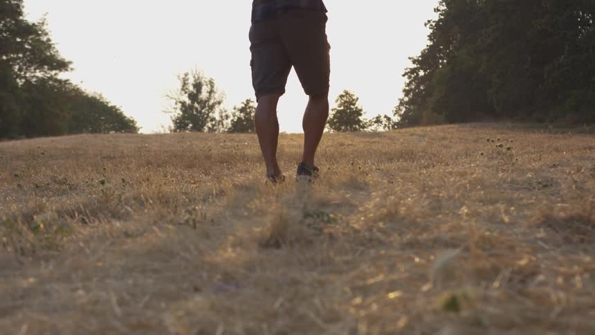 A man with muscular calves is wearing shorts tennis shoes a flannel shirt and a backpack as he hikes up a grass hill near a forest