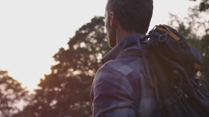 A man carrying a backpack looks ahead with the sun rising over trees in the distance