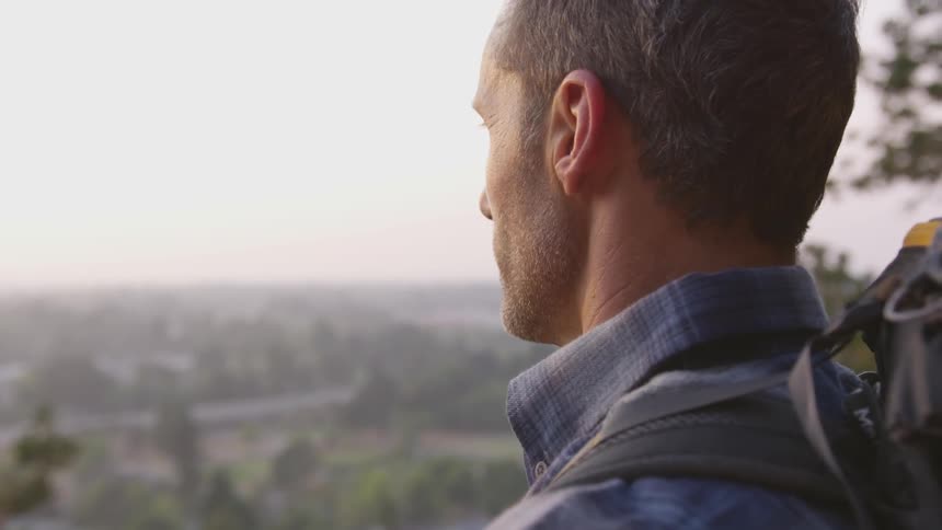 Man wearing blue striped shirt and backpack looks at the distant city at sunset