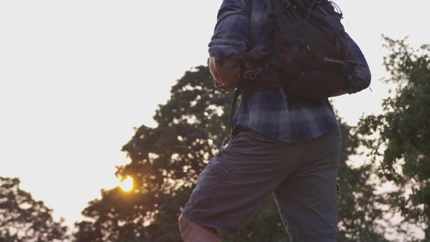 A man wearing a blue flannel shirt and gray shorts stands in a forested area while carrying a backpack on his back