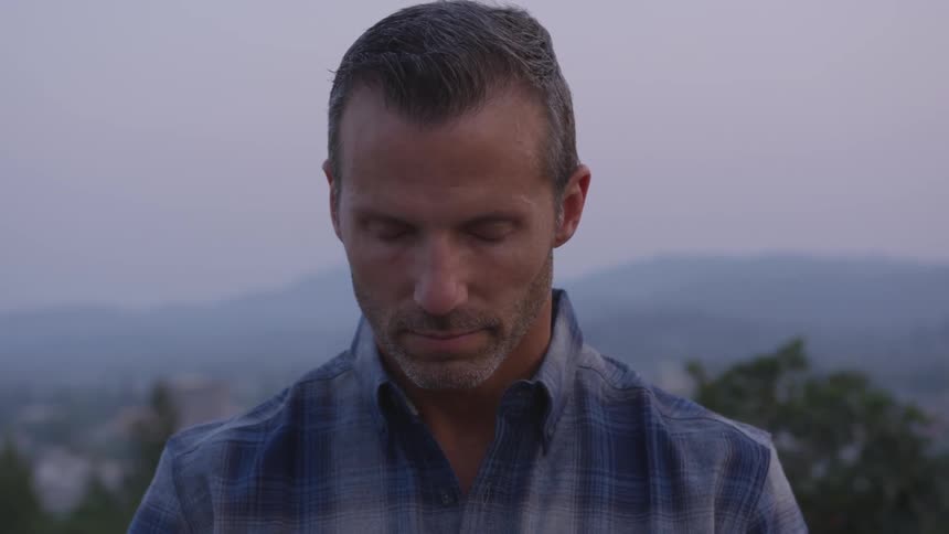 A man in a blue plaid shirt stands with eyes closed with the tops of trees and a mountainous landscape in the background