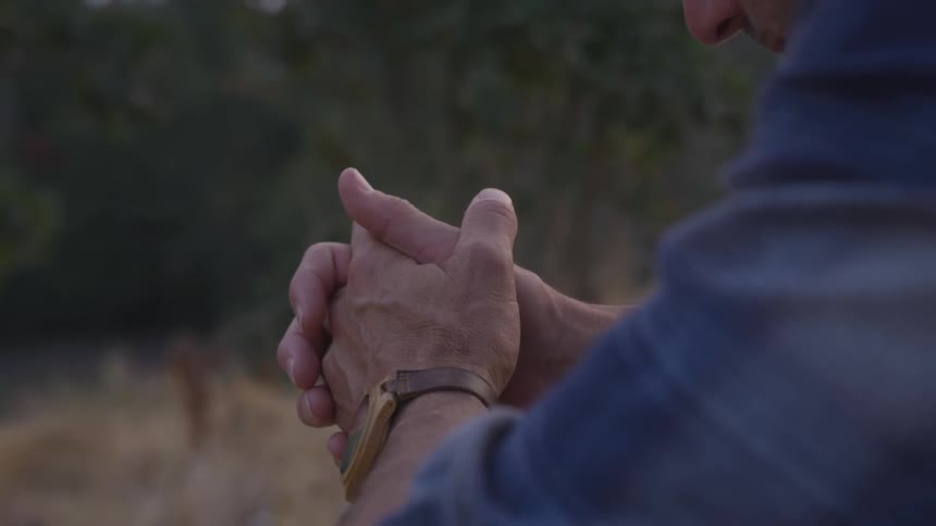 A man sits outside near trees with his hands clasped and looking down
