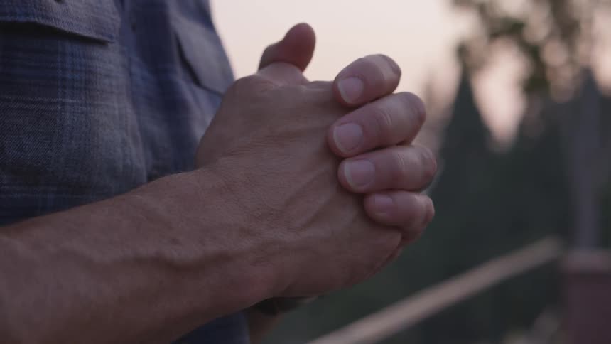 A man in a blue flannel shirt stands outside at dusk while holding his fist in his other hand