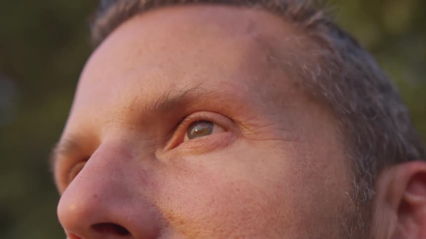 A man with gray hair is seen staring off into the distance while he is outside in a forested area