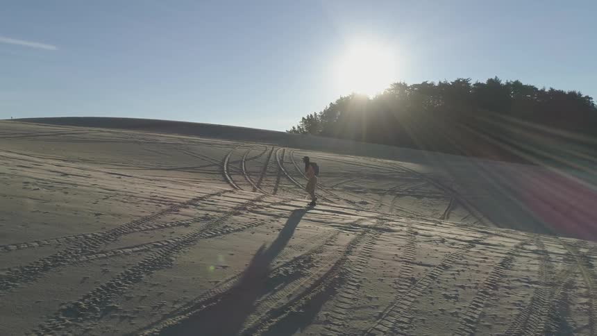 Person walks across tire track covered sand with trees in distance and sun on the horizon