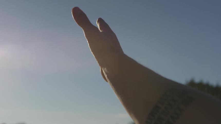 A man with a tattoo on his arm reaches his hand up towards the sunny blue sky