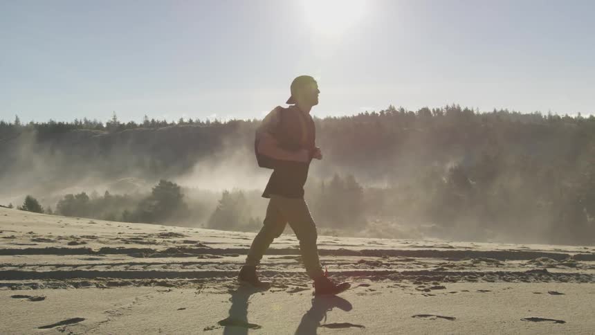 A man wearing a black t shirt khaki pants and boots carries a backpack as he walks along a sandy beach area with tire tracks imprinted in the sand