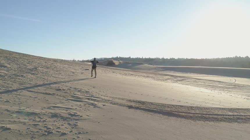 Man is walking with arm outstretched over a sandy hill with trees surrounding it on a sunny day
