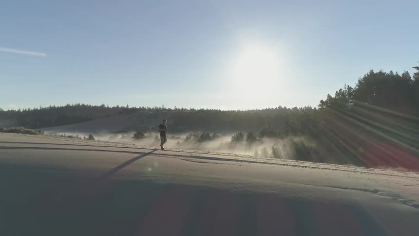 A man is hiking atop a sandy ridge that is adjacent to a dense forest with a layer of fog settled upon it