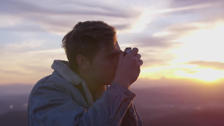 A man standing above a valley below lifts a camera up to take a photo at sunset