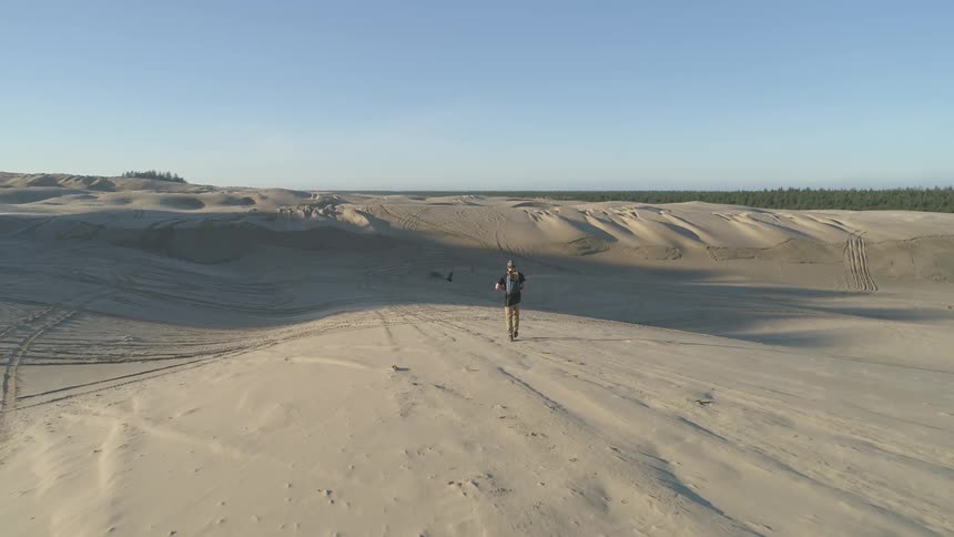 A man wearing a backpack walks across a sand dune with tire tracks on it