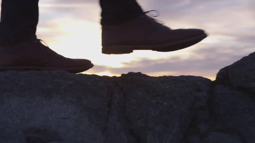 A man in brown shoes walks on top of a ridge of large rocks with a sunset in the background