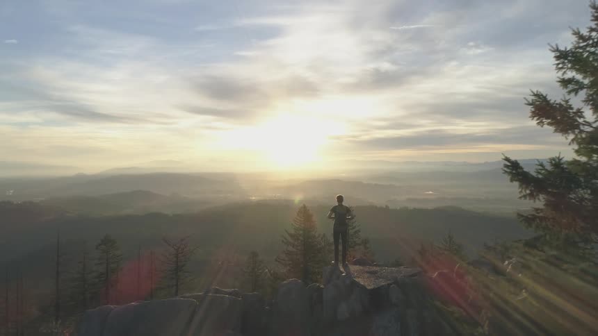 Man stands on mountain cliff overlooking forest at sunrise