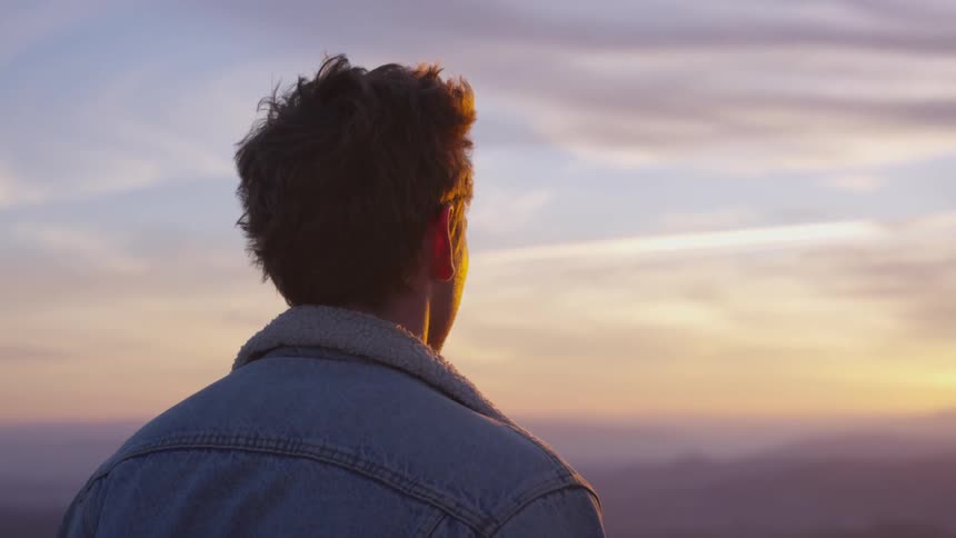 A man with messy hair and wearing a denim jacket stands atop a mountain and looks out over a sprawling valley