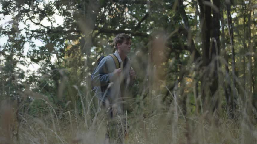 A young man wearing a backpack is hiking through some tall grass within a forest