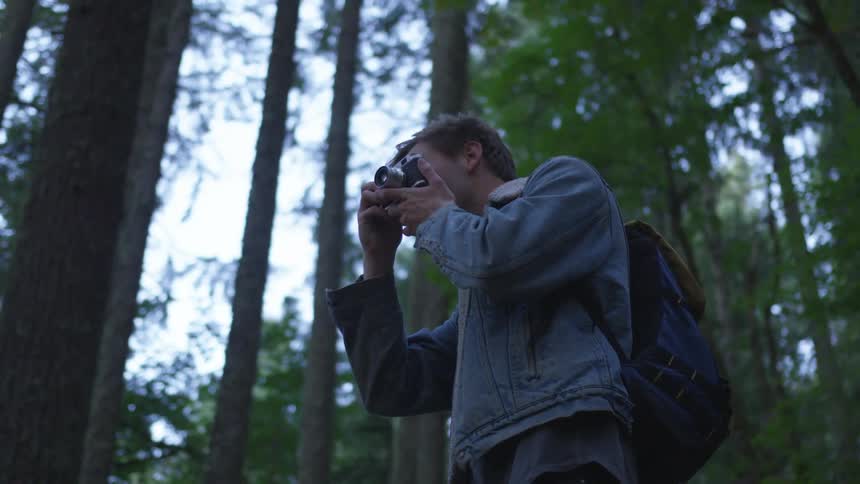 A male wearing a jean jacket and backpack is taking pictures with his camera among the tall trees in the woods