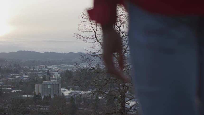 A person stands on a ridge overlooking buildings trees and hills at sunset