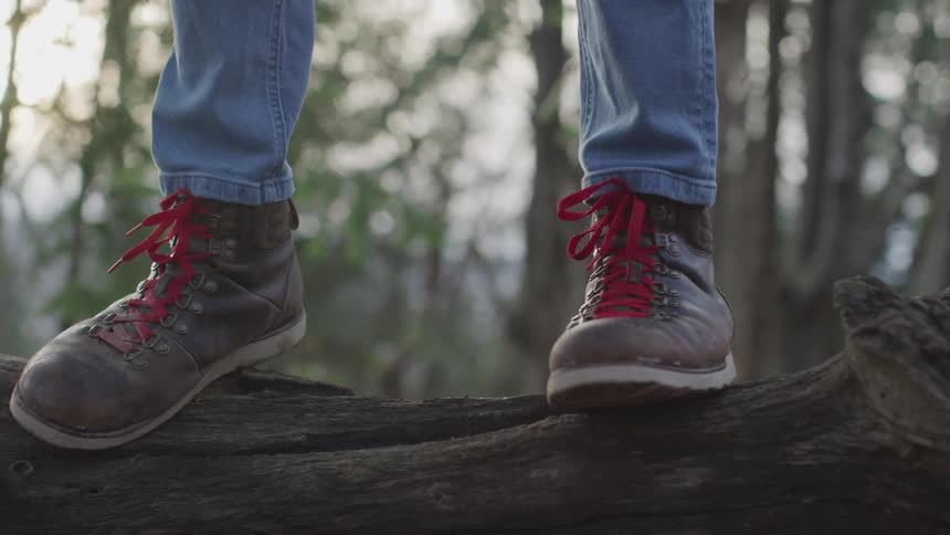 A man wearing blue jeans and brown leather boots steps atop a fallen log in a forests and tilts forward to maintain their balance