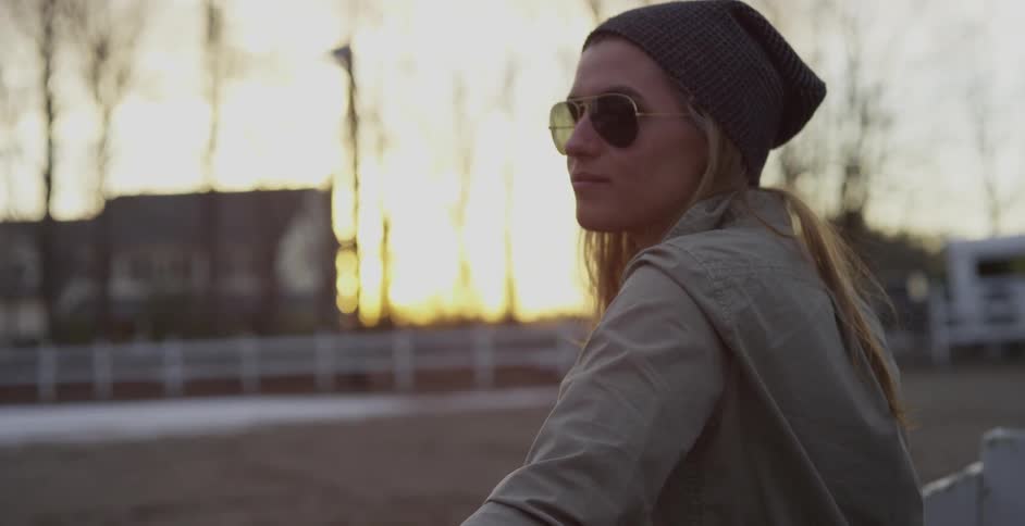 A blonde woman wearing a hat, standing next to a fence and looking at something in the distance