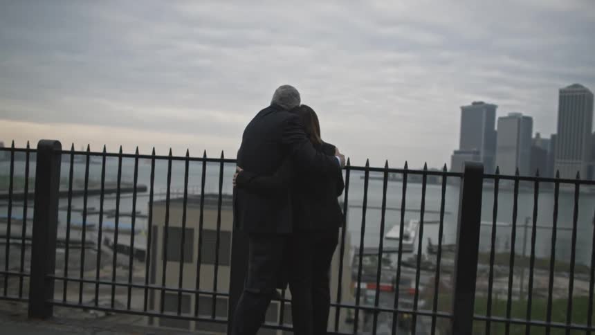 A couple embracing as they stand next to a fence and look over a body of water in an urban area