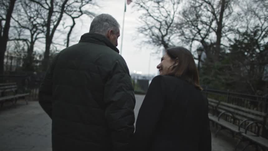 A couple talking, smiling and holding hands as they walk down a path lined with trees and benches