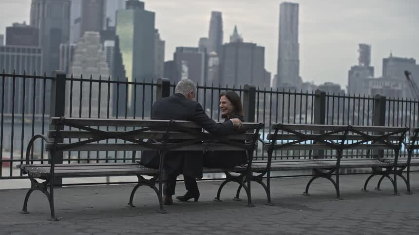A man and woman sitting on a bench and talking next to a fence and a body of water in an urban area