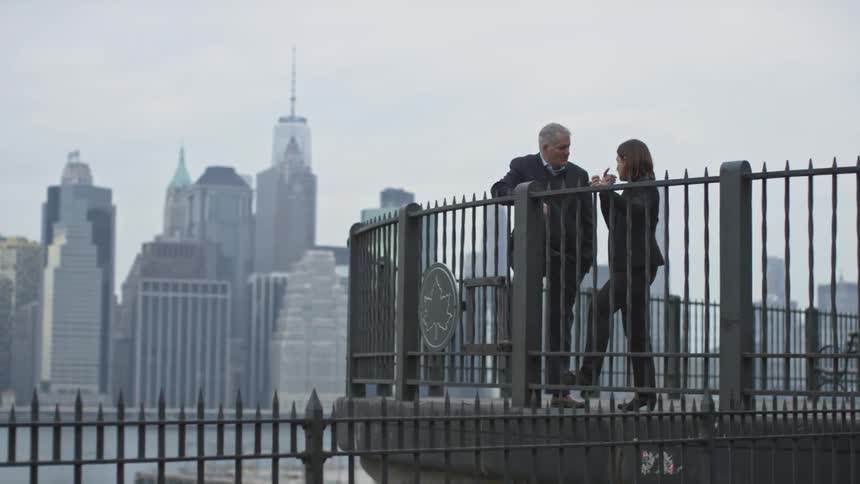 A young woman and a mature man having a conversation outdoors on a platform overlooking a river and skyline