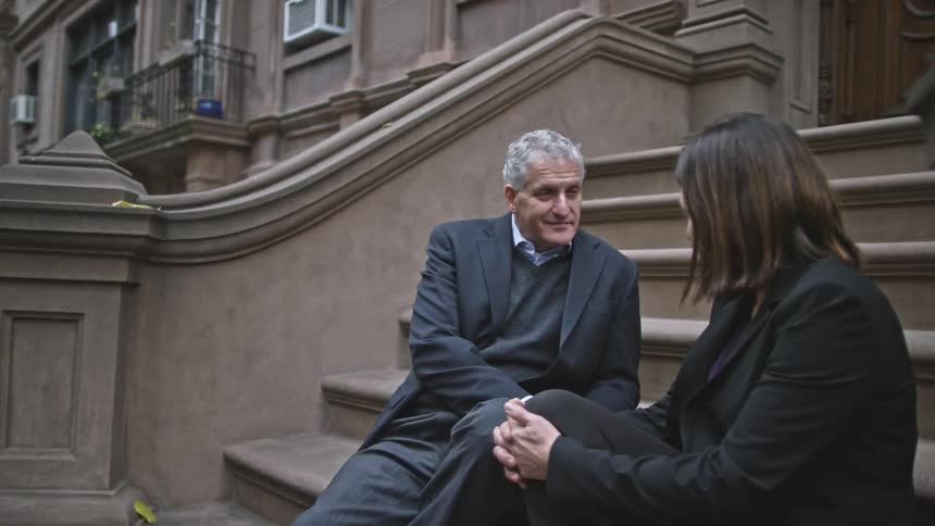 An older man and a younger woman sit talking on the steps of some upscale row houses