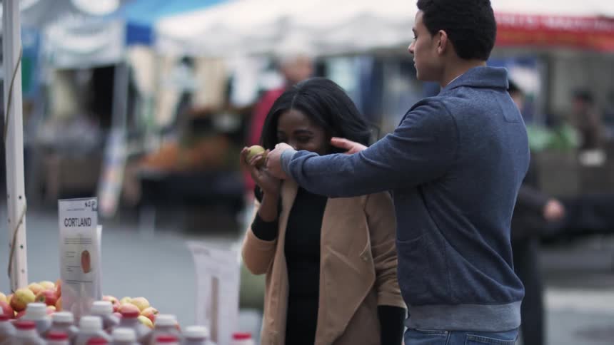 Man instructing a woman to smell some fruit from an outdoors fruit stand on a busy city street