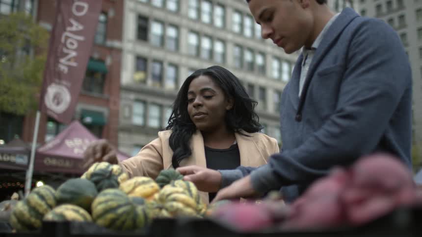 2 people discussing fruit at an outdoor fruit stand as others walk by on a city street