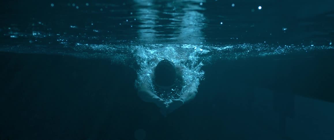 A dark underwater view of a man diving into water, as seen from the front