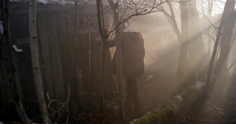 Man walks over to a wooden structure and enters on a cloudy day