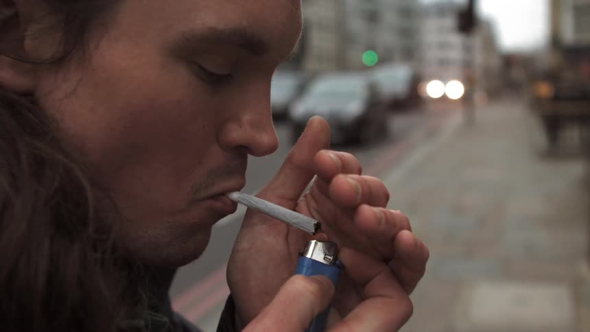 A younger man standing in an urban setting lights a homemade cigarette or possibly a joint