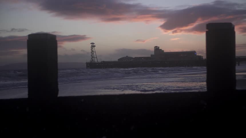 The ocean waves crash against a warehouse building and the shoreline