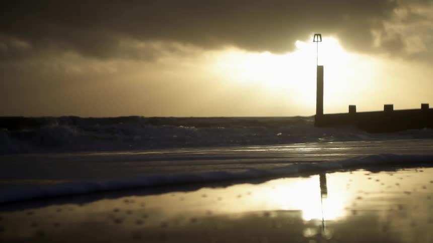 View of waves rolling onshore as the sun sets in a heavily clouded sky