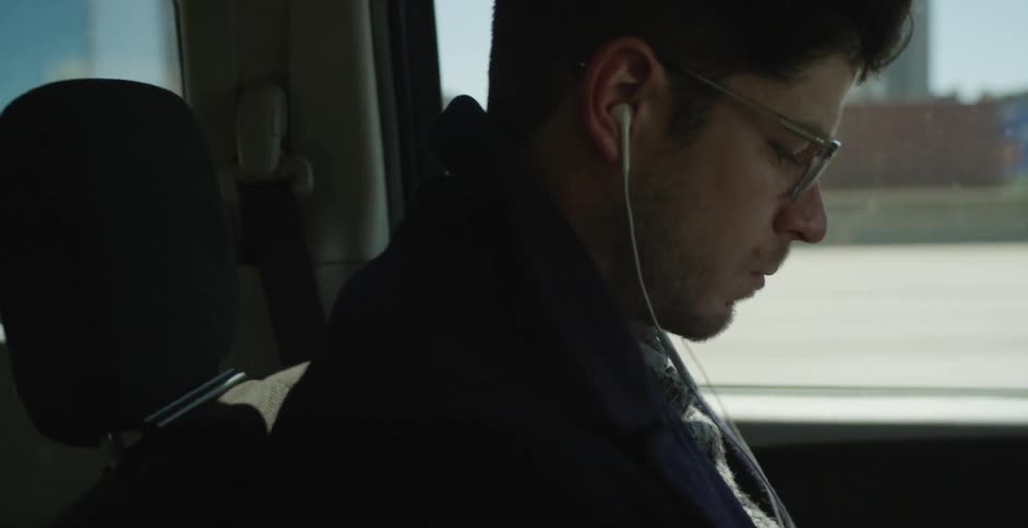 A man wearing earbuds sits in a van looking at something while commuting
