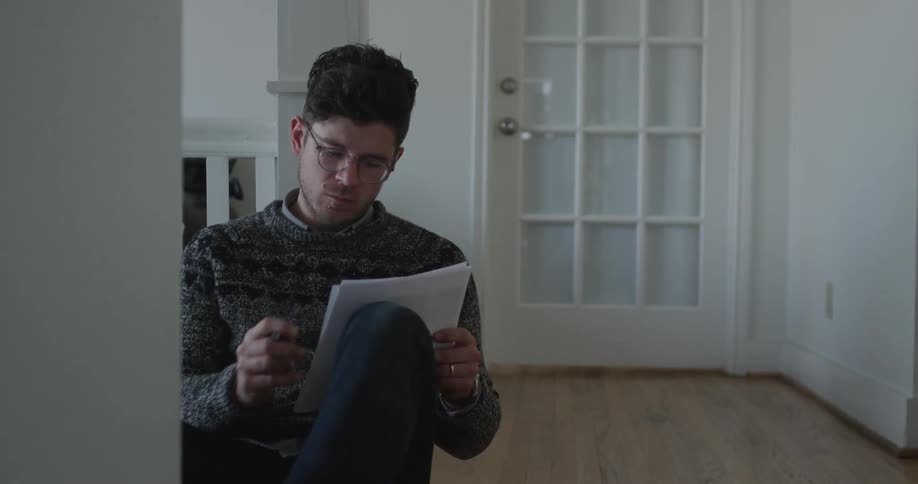A young man is seated on a wooden floor and flipping through sheets of paper