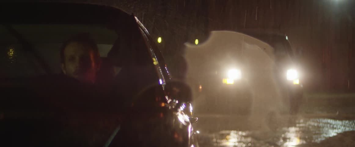 A man sits in a car at night in the rain as another car approaches