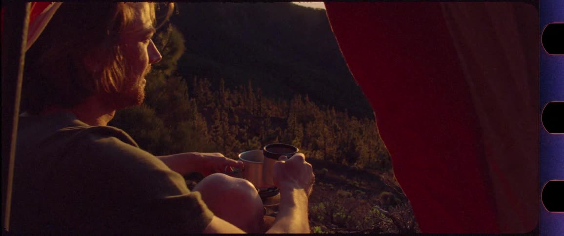 Man pouring coffee into his cup in front of his tent up in the mountains at sunrise