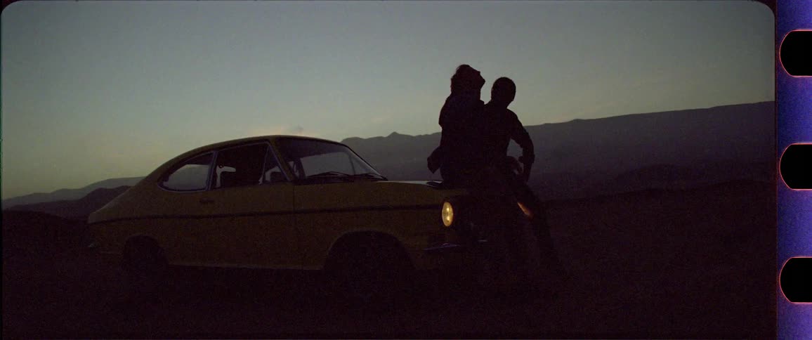 Two silhouettes sitting on a yellow car in front of a mountain desert region after sunset